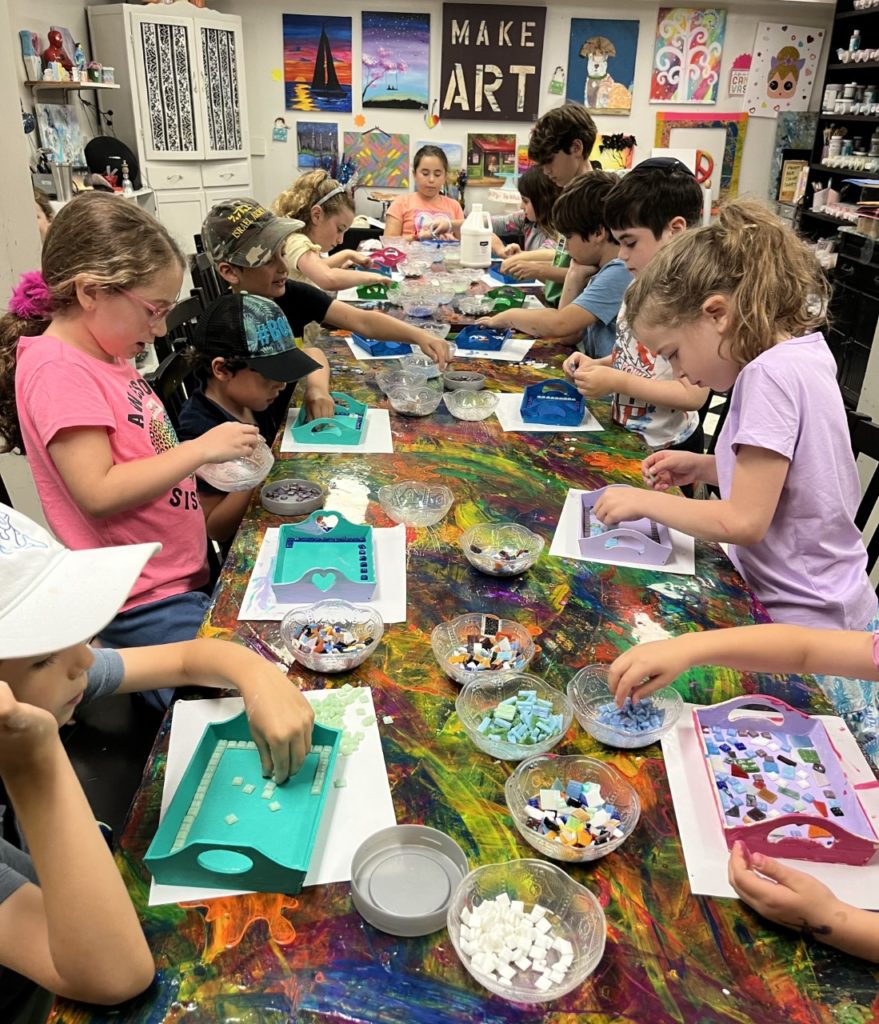 A table cluttered with colorful children's artwork. The table is covered with drawings and paintings featuring bright, and imaginative designs